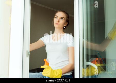 A cute adult female cleaning company worker wipes plastic windows in a house. The view from the outside. Stock Photo