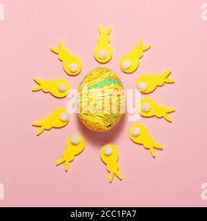 Yellow and green corduroy bunnies circling around an Easter egg in the center of the frame on a pink background. Flat lay top view Stock Photo