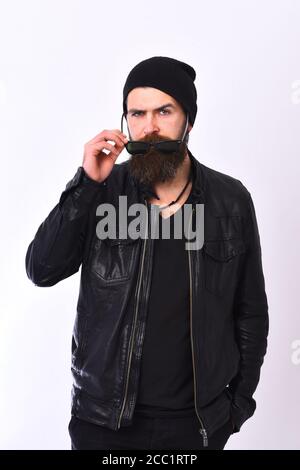 Rock and street life concept. Rock fan or gangster takes his glasses off. Man with serious face and brutal style isolated on white background. Guy with beard in black leather jacket and hat. Stock Photo