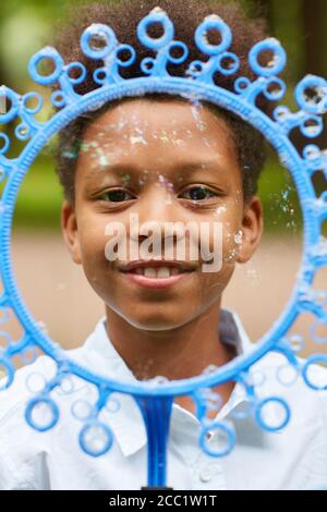Vertical close up portrait of smiling African-American boy looking at camera through big bubble wand while playing outdoors in park Stock Photo