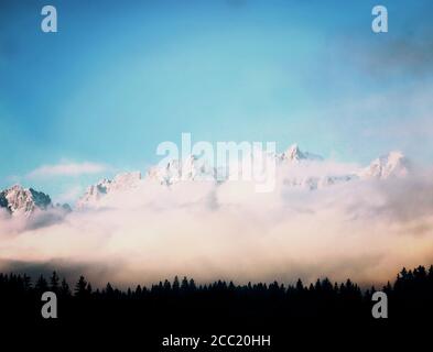 Beautiful view of Austrian Alps surrounded by lower clouds and towering over the fir tree forest Stock Photo