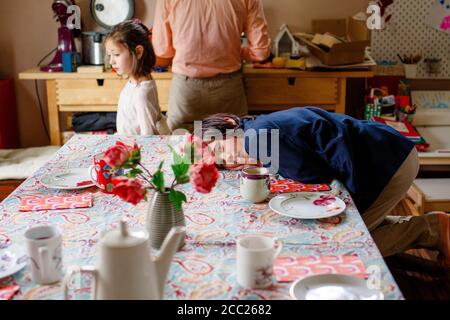 A boy in a suit lays across a table set for tea, family in background Stock Photo