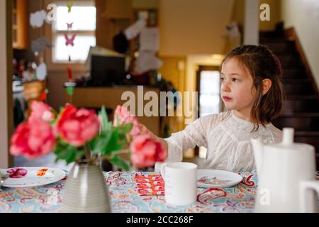 A small child in a lace dress sits alone at a table set for tea party Stock Photo