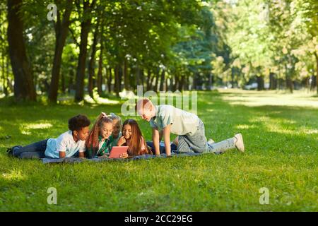Multi-ethnic group of kids using digital tablet while lying on green grass in park outdoors lit by sunlight, copy space Stock Photo