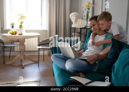 Son hugging working woman and focusing on laptop screen while sitting on sofa Stock Photo