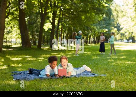 Wide angle portrait of two cheerful boys using digital tablet while lying on green grass in park outdoors lit by sunlight, copy space Stock Photo