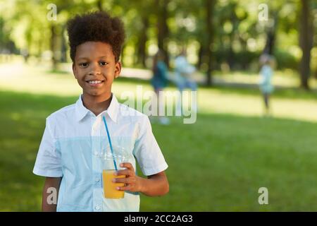 Waist up portrait of smiling African-American boy holding orange juice while standing in green park outdoors, copy space Stock Photo