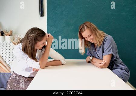 Two beautiful women laugh. Female businesswoman sit at table in workplace and talk, have fun Stock Photo