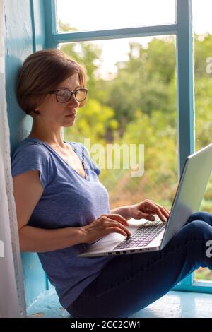 Woman freelancer sitting on a windowsill working on a laptop Stock Photo