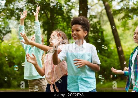 Waist up portrait of African-American boy and group of carefree children running in park while playing with bubbles outdoors Stock Photo