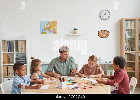 High angle view at male teacher working with multi-ethnic group of children drawing pictures during art class in school or development center, copy sp Stock Photo