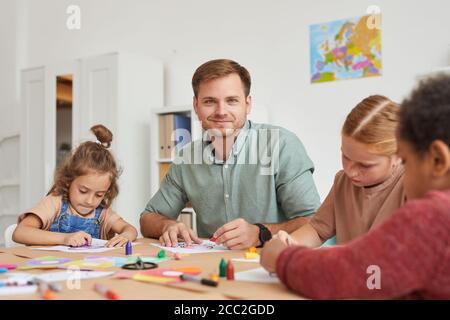 Portrait of male teacher smiling at camera while working with multi-ethnic group of children drawing pictures during art class in school or developmen Stock Photo