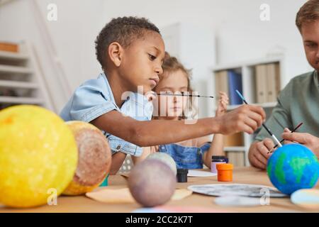 Side view portrait of cute African-American boy painting planet model while enjoying art and craft lesson in school or development center, copy space Stock Photo