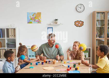 Portrait of smiling male teacher working with multi-ethnic group of children painting planet models in art and craft lesson, copy space Stock Photo