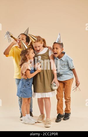 Vertical full length portrait of diverse group of children wearing party hats screaming to microphone while standing against plain background in studi Stock Photo