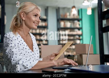 Side view of happy succesful business owner holding an envelope or parcel, using laptop and smiling while sitting in her craft pottery shop with handmade ceramic products. Small business, retailing Stock Photo