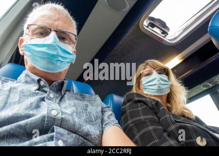 Middle aged man and woman travelling on public transport during UK Covid-19 epidemic wearing surgical masks. Looking down at the camera. Stock Photo