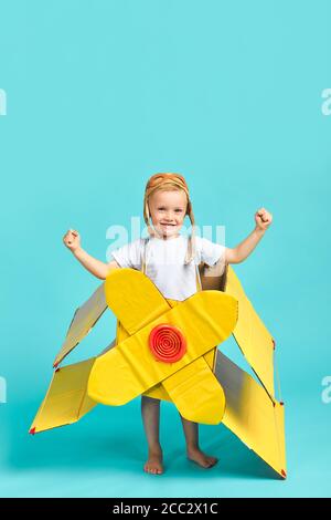 Playful boy wearing white t-shirt and toy airplane, helmet on head stand happily lokking at camer in isolated blue studio. Sky, pilot, aviation concep Stock Photo