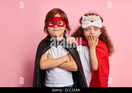 Two cute kids wearing hero cloaks and masks stand isolated over pink background, togetherness, children team. Suprehero concept. Surprised girl closed Stock Photo