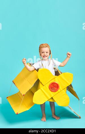 Caucasian child in yellow toy airplane show muscles at camera, child's power. Imagination, fantasy, dreams. Portrait of boy isolated over blue backgro Stock Photo