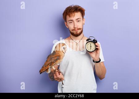 Sleepy unhappy man with an owl on his arm holds alarm clock after waking up in the morning.close up portrait, lifestyle, free time, spare time Stock Photo