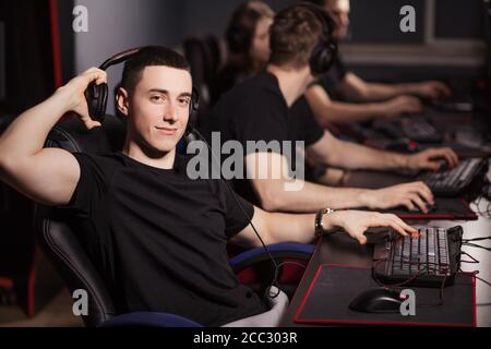 Satisfied smart gamer in black t-shirt sitting behind the computer monitor and looking at camera in a gaming club or internet cafe with the other peop Stock Photo