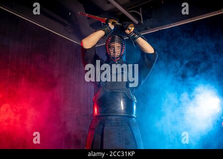 caucasian male kendo fighter in traditional Japanese style of clothing, protective armour, using shinai. practicing fight isolated over smoky room Stock Photo
