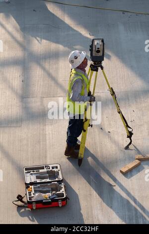 Construction worker wearing safety gear and face covering uses tripod-mounted transit on parking garage job site near downtown Austin, Texas. Stock Photo