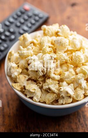 Sweet tasty popcorn in bowl and TV remote control on wooden table. Stock Photo