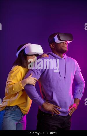 Young interracial couple having first experience of using virtual reality headset, standing joyful and impressed in studio with violet background Stock Photo
