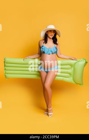 young pregnant woman in swimsuit posing with inflatable mattress on yellow Stock Photo