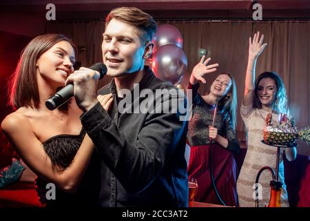 lovely young caucasian couple spend free time, celebrating in karaoke bar, man and woman in black wear singing together in microphone Stock Photo