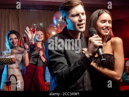 beautiful woman and man, couple have free time in karaoke club, happy woman and man in black wear singing in microphone with friends in the background Stock Photo