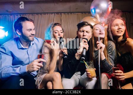 young women and men clubbers spend time in karaoke bar, have fun singing in microphone. leisure, celebration, party concept Stock Photo