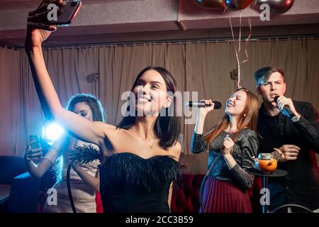 lady in the long black dress karaoke young and beautiful brunette take selfie with friends in bar, friends with balloons in background, smoky space Stock Photo karaoke night dress code