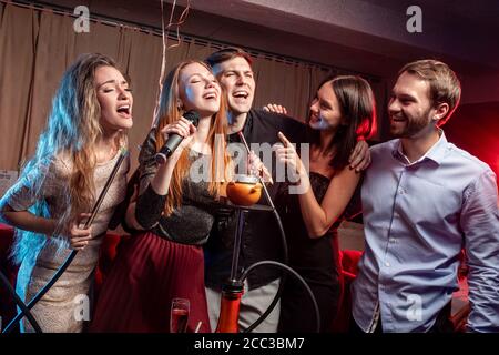 young women and men clubbers spend time in karaoke bar, have fun singing in microphone. leisure, celebration, party concept Stock Photo