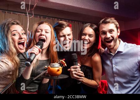 young caucasian students gathered to sing in karaoke bar, having fun together, karaoke and smoking hookah isolated in room Stock Photo