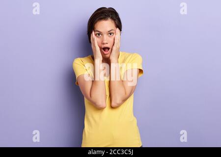 attractive girl with palms on her cheeks being in panic, close up portrait, isolated blue background, studio shot. feeling and emotion. oh. my God Stock Photo