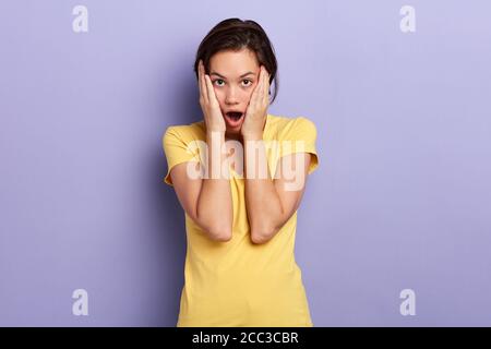 emotional girl touching her cheeks, expresses amazement, has mouth wide opened, can not believe in shocking news, isolated over blue. lifestyle, facia Stock Photo