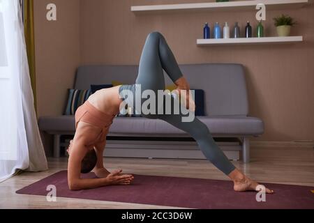 Athletic and sportive girl practice yoga bridge at home in living room, one leg is raised above another. Stock Photo