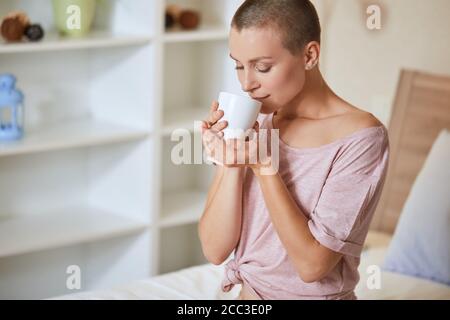 Young beautiful girl with short hair sit on bed in light room and enjoy drinking tea or coffee at morning at home. Stock Photo