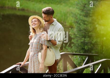 Portrait of romantic adult couple smiling happily while walking across wooden bridge by lake in rustic country scenery, copy space Stock Photo