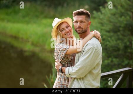 Waist up portrait of romantic adult couple embracing and looking at camera while standing on wooden bridge by lake in rustic country scenery, copy spa Stock Photo