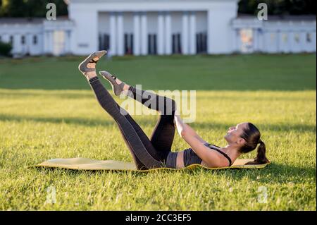 Beautiful young woman lying on a yellow mattress, pose while wearing a tight sports outfit in the park doing pilates or yoga, single leg stretch inter Stock Photo