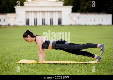 Beautiful young woman lying on a yellow mattress, pose while wearing a tight sports outfit in the park doing pilates or yoga, leg pull superior exerci Stock Photo