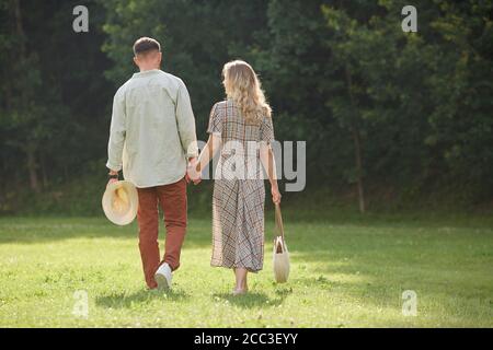 Back view portrait of romantic adult couple holding hands while walking on green grass in nature scenery, copy space Stock Photo