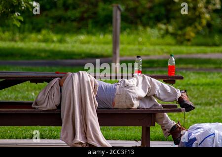A homeless caucasian man is sleeping on a picnic table in a shaded area of a park. He is covering his head with a dirty blanket. His belongings are in Stock Photo