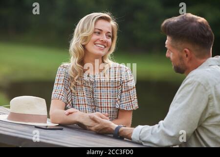 Portrait of romantic couple holding hands and looking at each other with love while sitting at outdoor table by lake, copy space Stock Photo