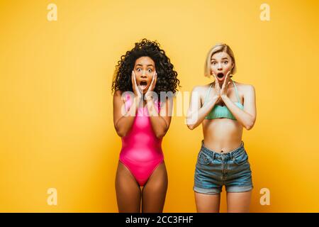 shocked multicultural women in summer outfit touching faces on yellow Stock Photo