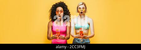 horizontal image of shocked interracial women in summer outfit holding cocktails and looking at camera isolated on yellow Stock Photo